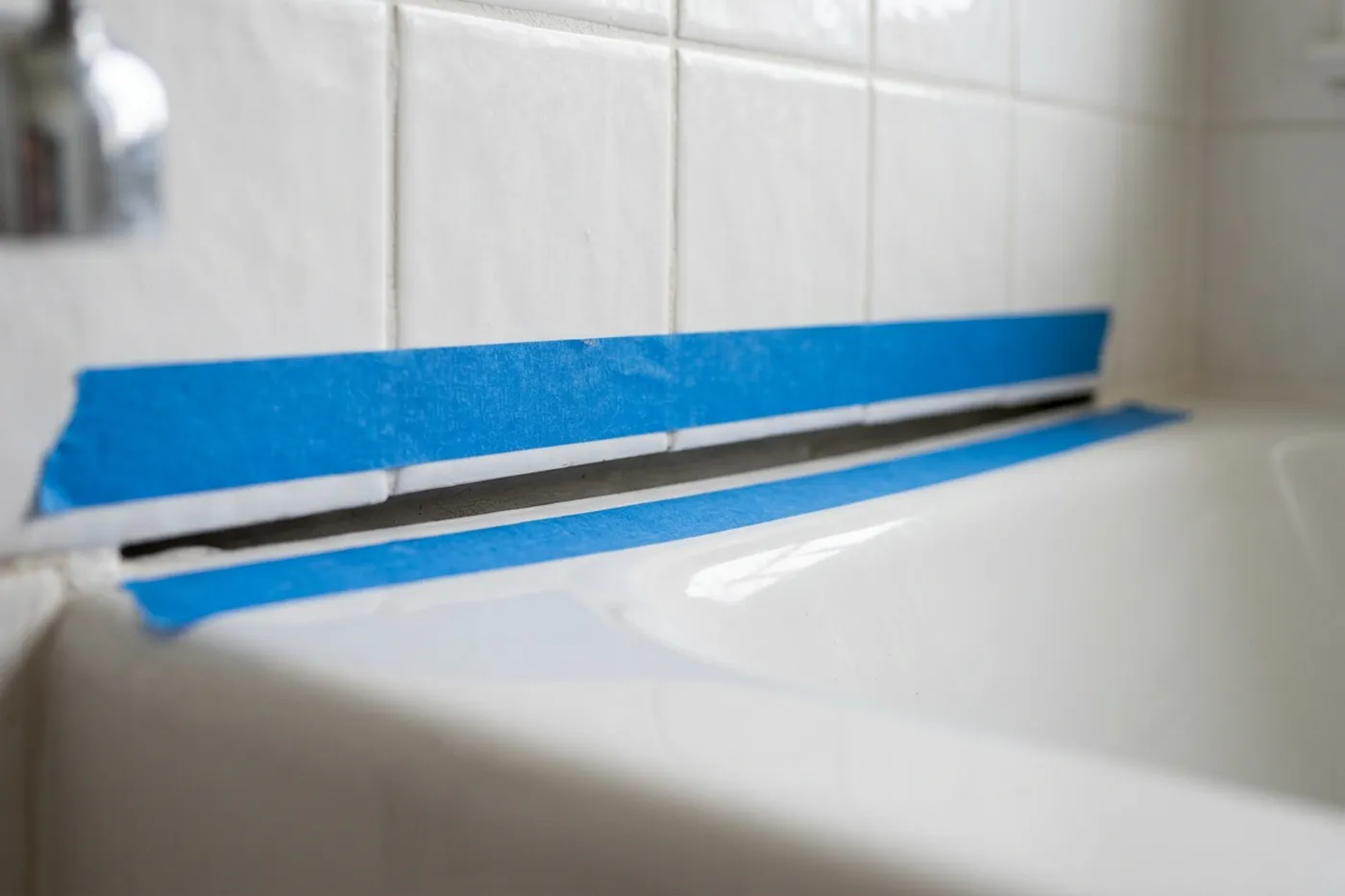 Two straight parallel strips of blue low-tack masking tape run either side of a bath joint, leaving a clean channel between them ready for sealing