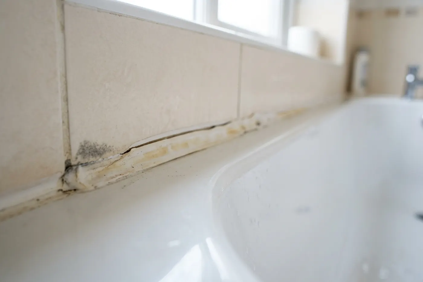 An ageing white silicone bead along a bath to tile joint photographed at a low angle along its length, showing where it has yellowed, split and lifted away from the tile